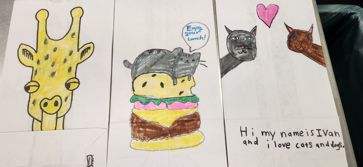 Art on student-decorated bags for the meals on wheels program. One has a giraffe, another has a cat in a hamburger. Yet another has a turtle and the phrase "I like turtles, do you?"