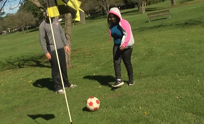 A high school student kicking a soccer ball into a golf hole, with a college student close at hand.