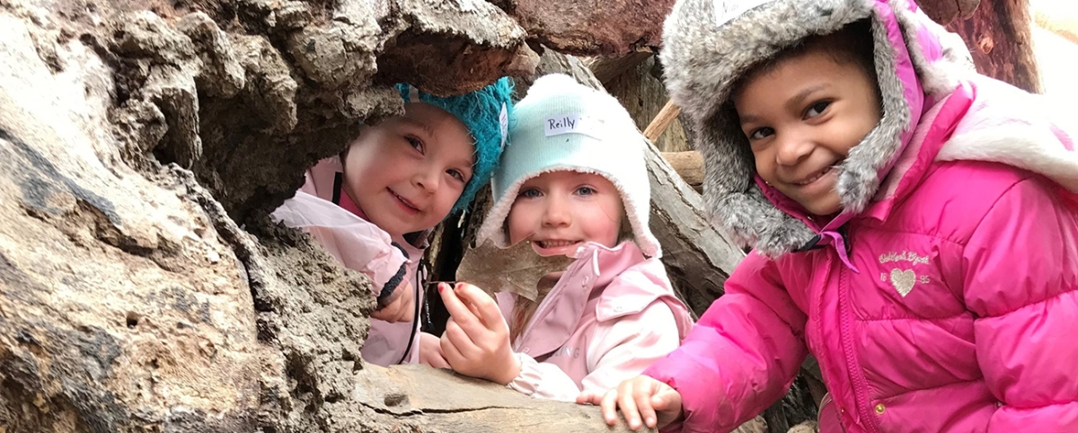 Three pre-kindergarten students smiling wearing winter gear and standing under a tree.