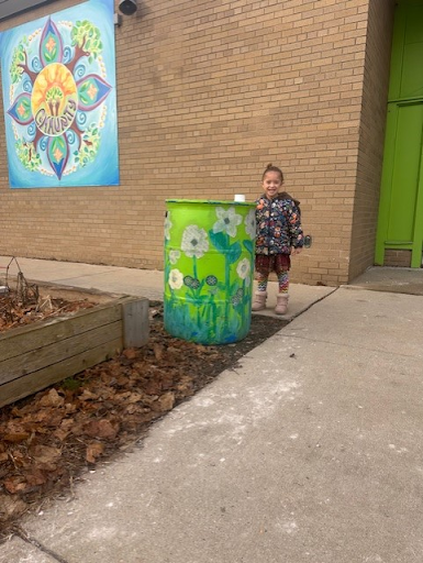 A kindergarten student stands proudly next to a newly-installed rain barrel at Parkside Elementary. The side of the rain barrel has been painted by students with a colorful flower.