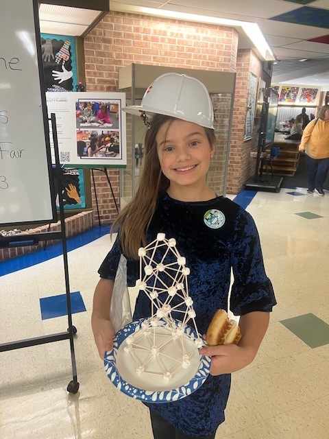 A student wearing a hard hat shows off their work at the STEAM fair (a structure made with marshmallows and toothpicks)