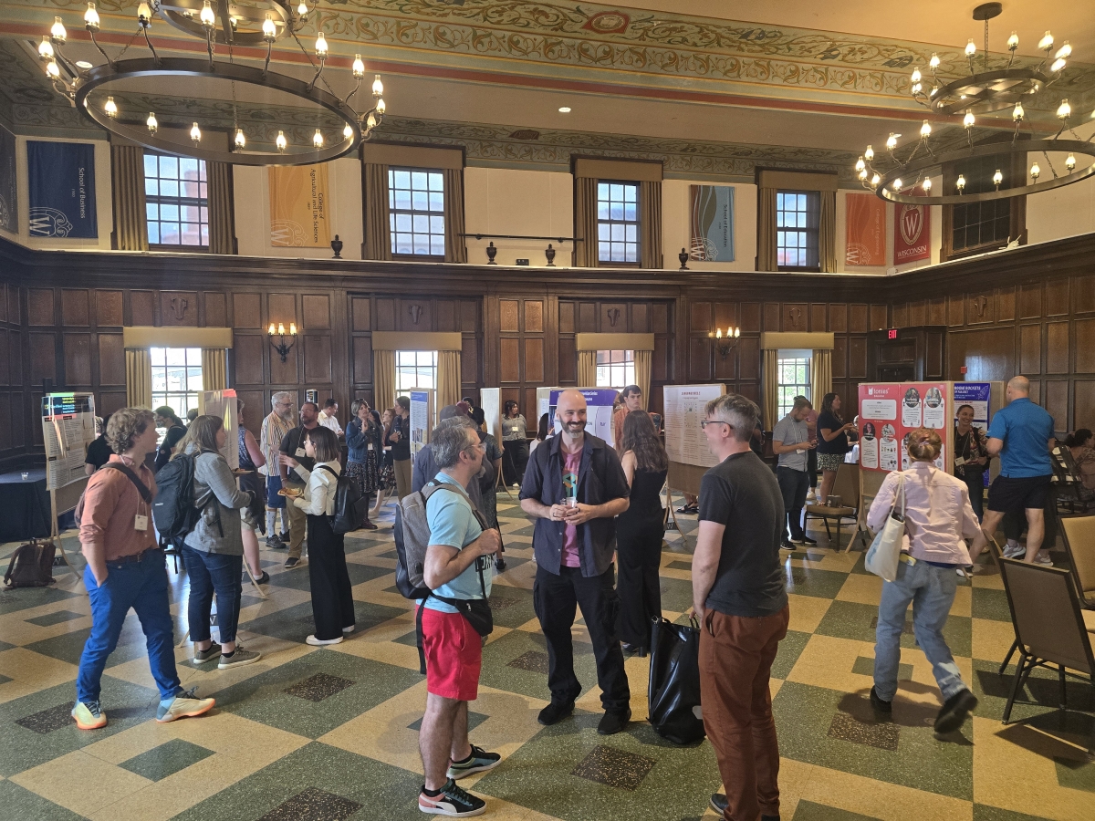 Poster Presentation session on the evening of July 18th in a large room teeming with people. There are posters in stations throughout the room, and participants can be seen congregating in front of the posters and having animated conversations with one another. 