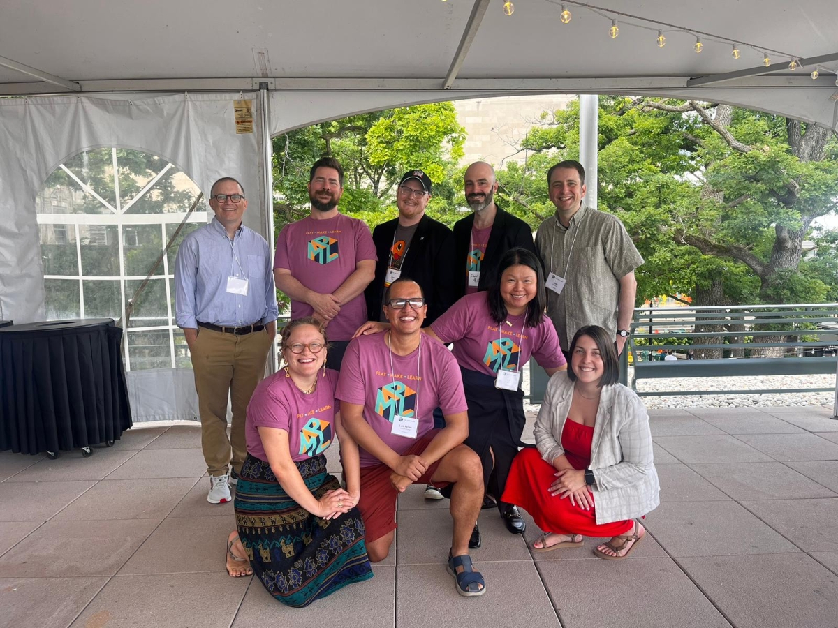 A group of 9 smiling people, many wearing a pink play make learn conference t-shirt, poses happily together in an outdoor event tent