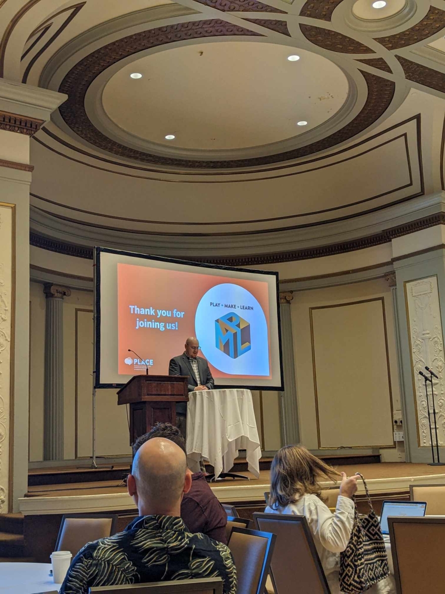 Chris Baker, Public Library Consultant, Games and Learning Consultant, welcomes attendees. He's seen standing behind a podium on a stage in front of a large screen displaying the play make learn conference logo and the words "thank you for joining us"